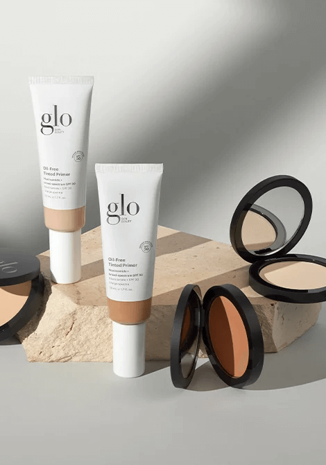 Two glo Oil-Free Tinted Primer tubes (SPF 30) and three open powder compacts on a stone slab, soft neutral lighting.