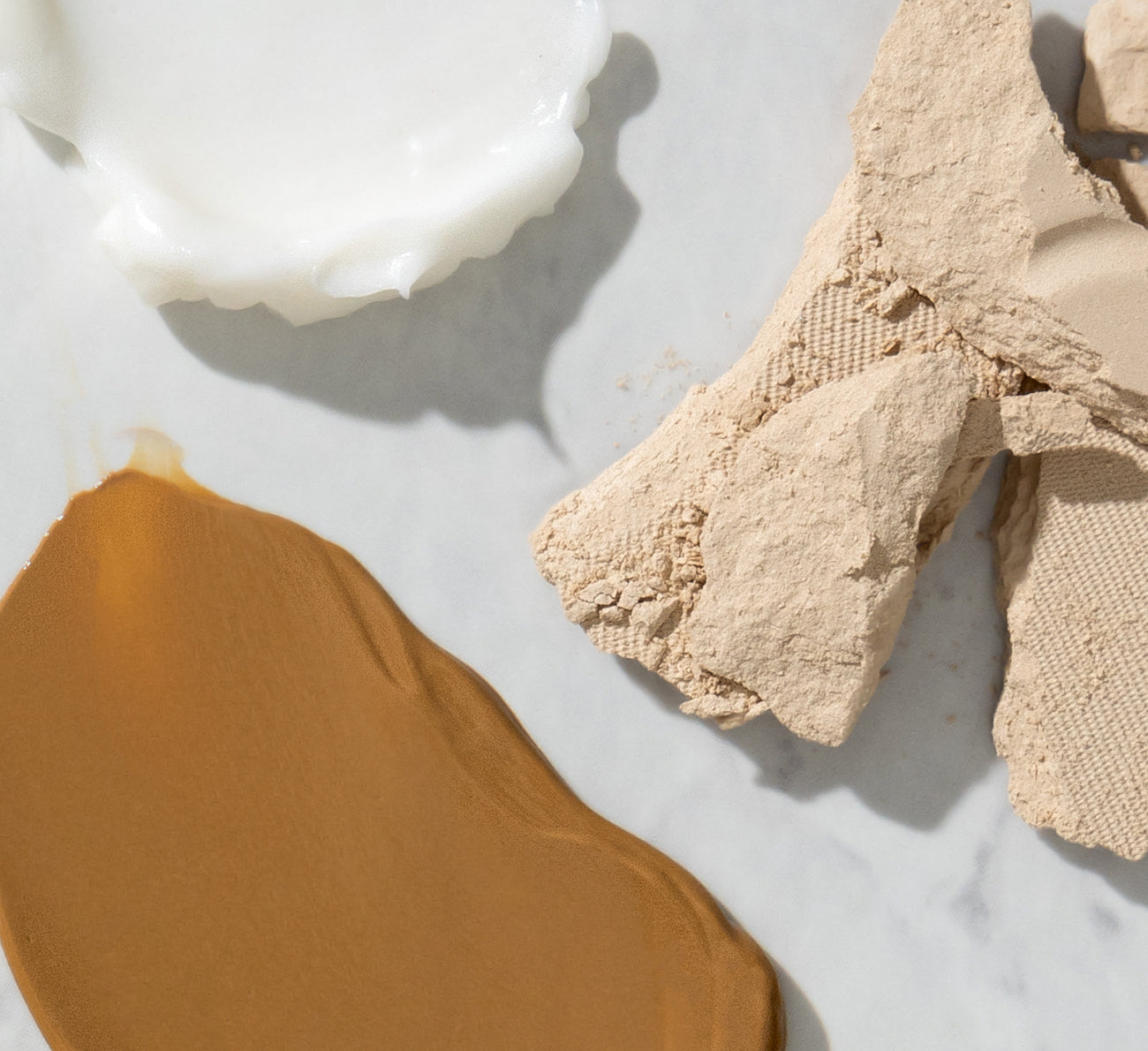 Swatches of tan liquid foundation, white cream moisturizer, and crumbled beige pressed powder on a marble surface.