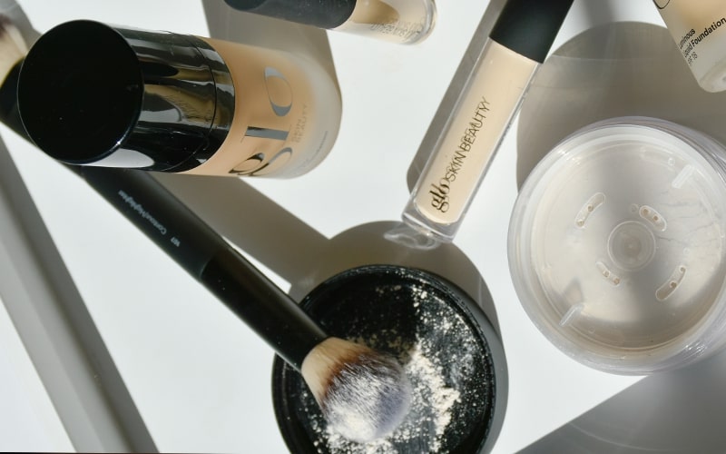 Flat lay of foundation bottles, concealer tube, loose powder, and a powder brush dusted with powder
