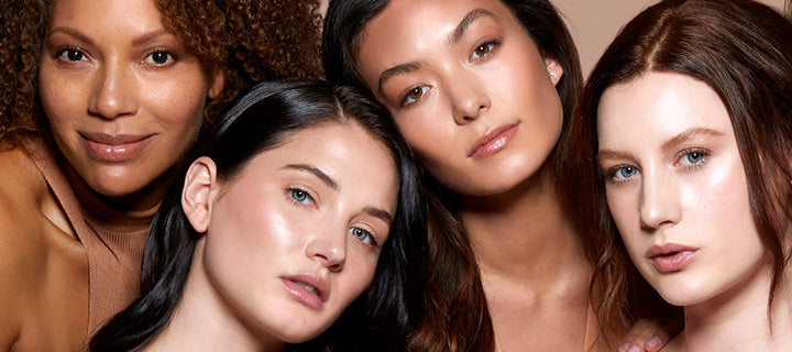 Four models, close-up faces showing diverse skin tones and dewy makeup, heads angled together looking at camera.
