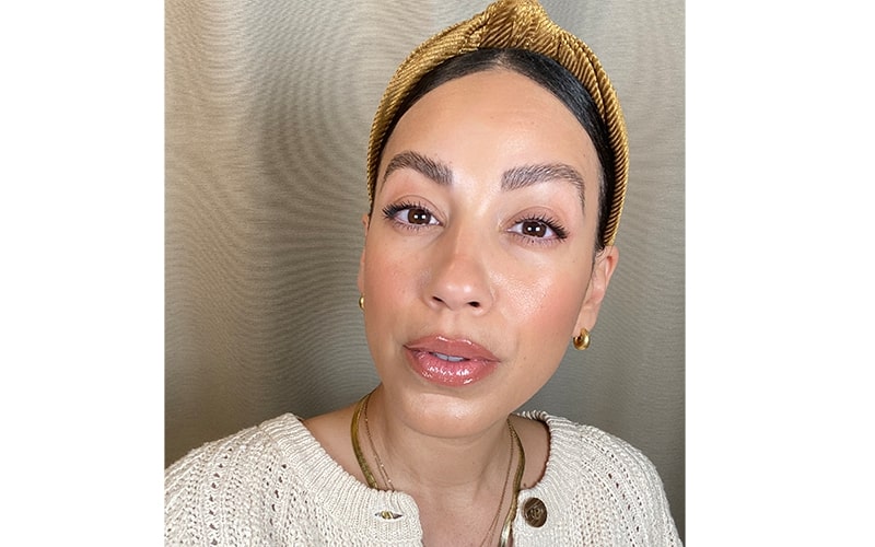 Close-up portrait of a person wearing a gold headband and earrings, with glossy nude contoured lips and natural brows.