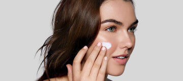 Young woman applying facial cream to her cheek, close-up showing hand and dab of product.