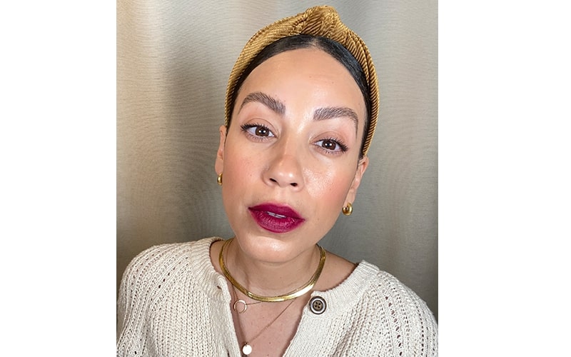 Close-up headshot of a woman wearing plum lipstick, gold headband, hoop earrings, and a cream knit sweater.
