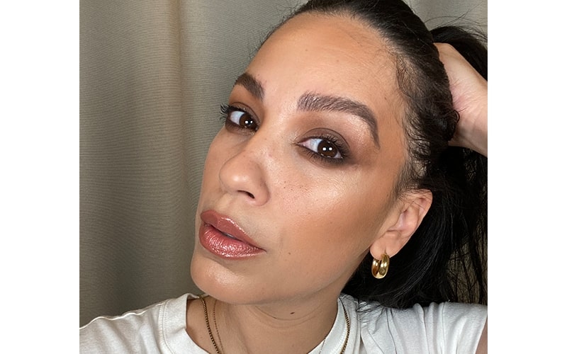 Woman wearing brown grunge smokey eye makeup, glossy lips, gold hoop earring, hair pulled back.