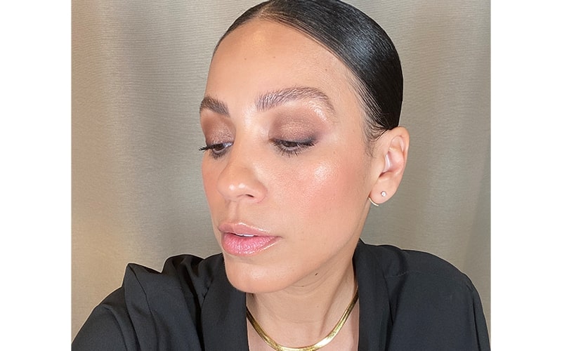 Close-up of a woman with brown smokey eye makeup, dewy skin, slicked-back hair and a gold necklace.