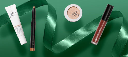 Four glo cosmetic products — cream tube, pencil, compact and lip gloss — arranged on green satin ribbon background.