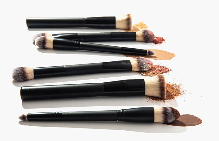 Five black-handled makeup brushes laid horizontally with scattered powder and foundation swatches.