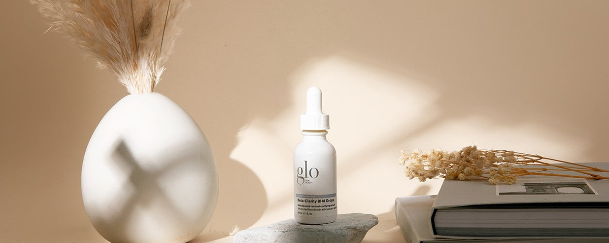 White dropper bottle labeled glo, Beta-Clarity BHA Drops on a stone beside dried flowers and stacked books in warm beige light.
