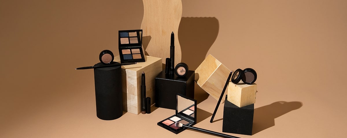 Assorted neutral eyeshadow palettes, pencils, and brushes arranged on wood and black blocks, with a 'glo' compact.