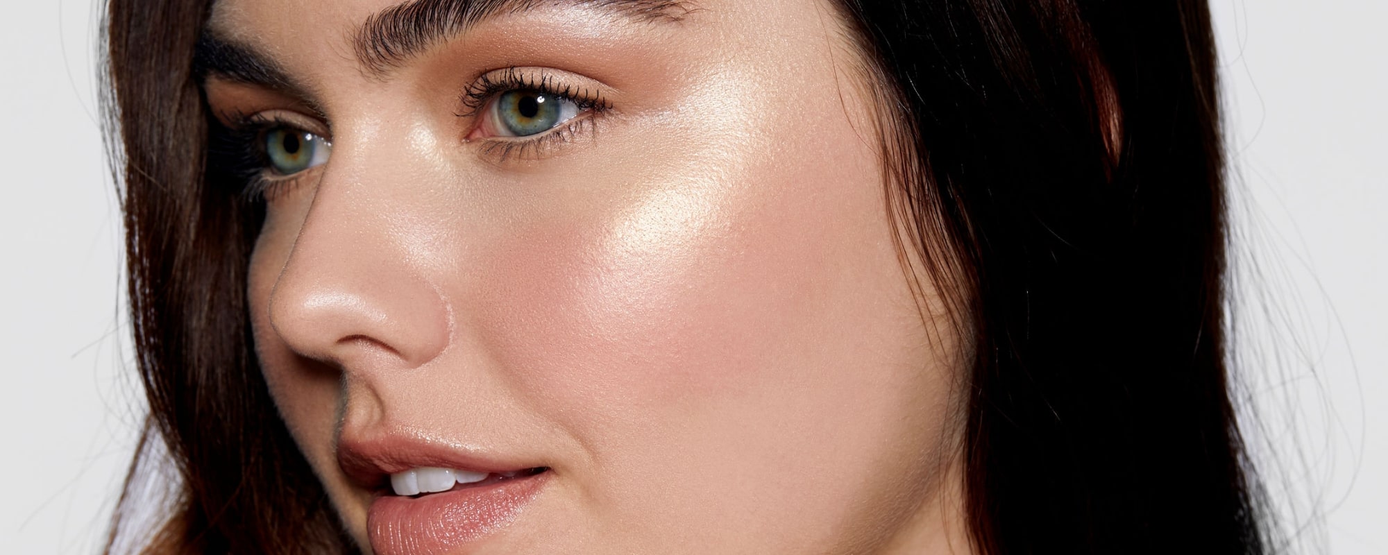 Close-up of a woman's face showing luminous highlighter on the cheekbone and natural makeup.