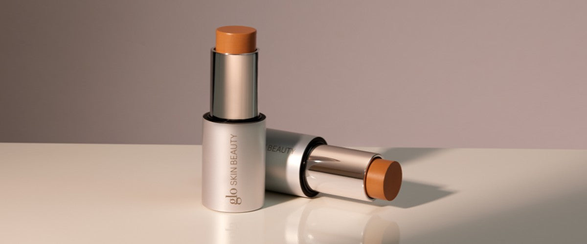 Two cream contour sticks in silver tubes on a reflective surface, labeled "glo SKIN BEAUTY".