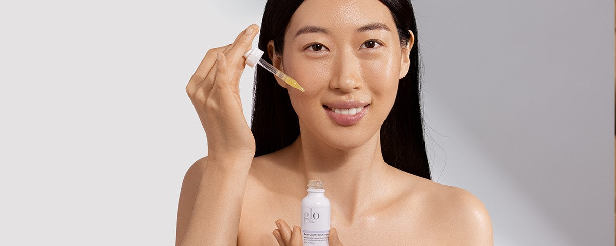 Woman smiling, holding a dropper applying facial serum above a bottle labeled 'glo'.