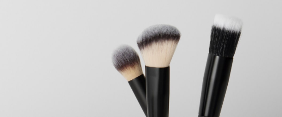 Three black-handled makeup brushes—angled blending, rounded powder, and flat foundation—on a pale gray background.