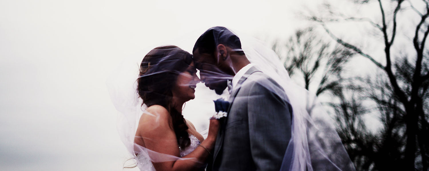 Bride and groom touching foreheads under the bride's veil, outdoors with bare winter trees behind them