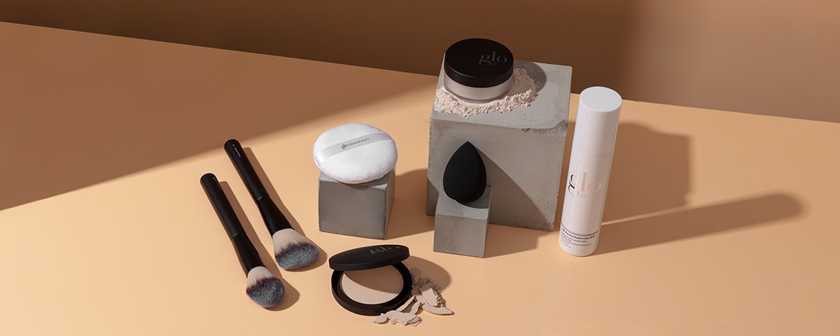 Makeup setting powders and tools on beige surface: loose powder jar labeled "glo", pressed compact, puff, sponge, and three brushes.