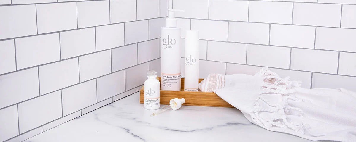 Skincare bottles labeled 'glo' on a marble bathroom counter with wood tray and white fringe towel