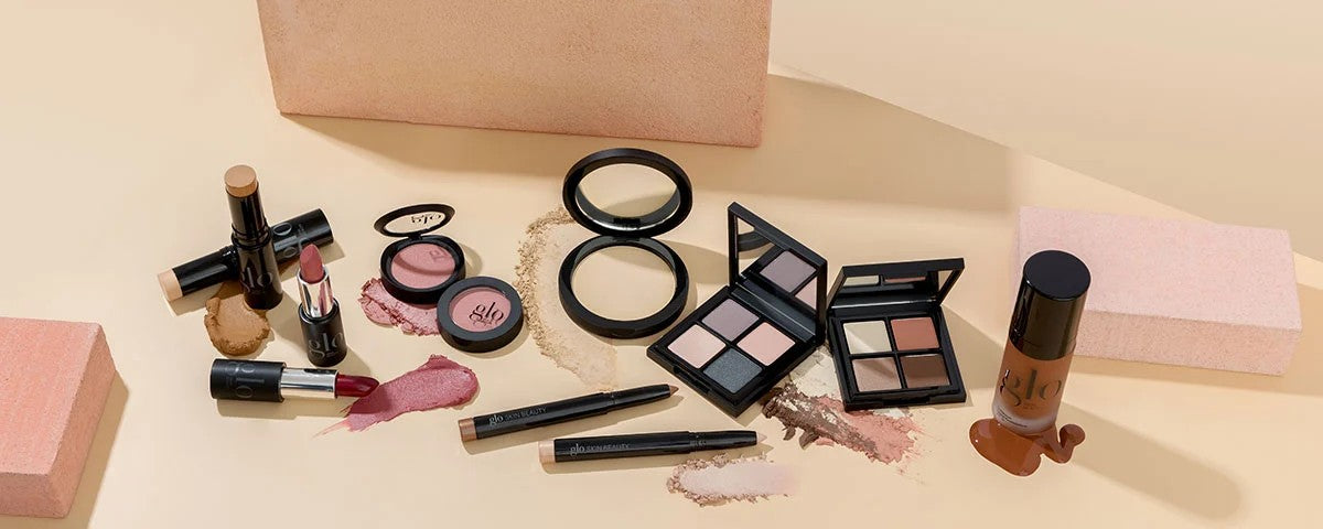 Assorted glo skin beauty cosmetics: lipsticks, cream foundation, blush compacts, eyeshadow palettes, and eyeliners on beige surface.