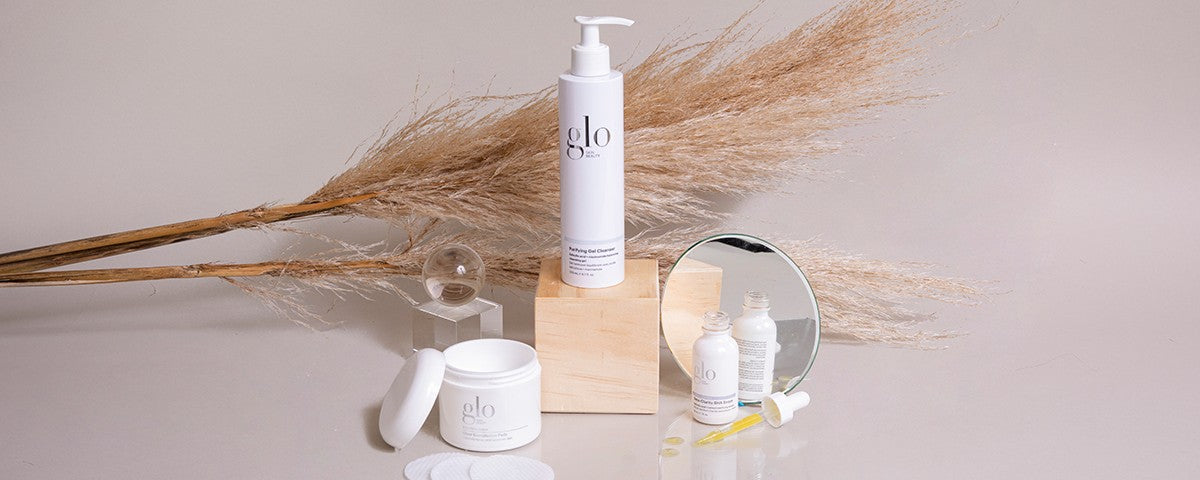 Glo skincare display: pump bottle labeled 'glo Purifying Gel Cleanser', jar, dropper bottle, mirror and pampas grass backdrop.