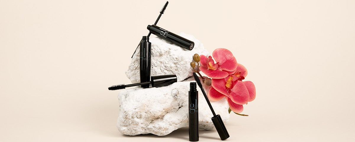 Three black mascara tubes on white rocks beside a pink orchid, with "gl" visible on one tube.
