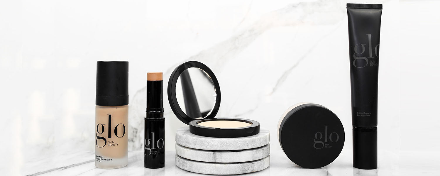 Assortment of glo SKIN BEAUTY foundation products—bottle, stick, cream compact and tube on a marble surface.