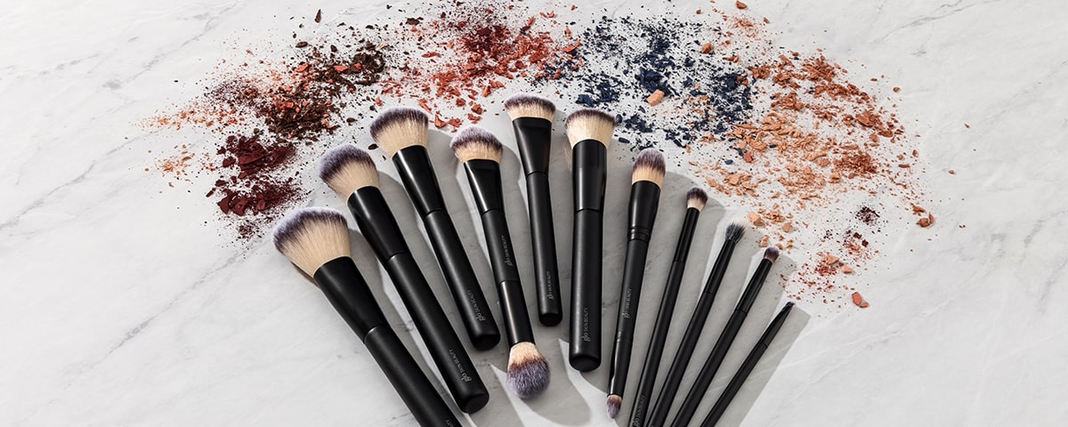 Twelve black-handled makeup brushes fanned on marble with scattered crushed eyeshadow and powder.