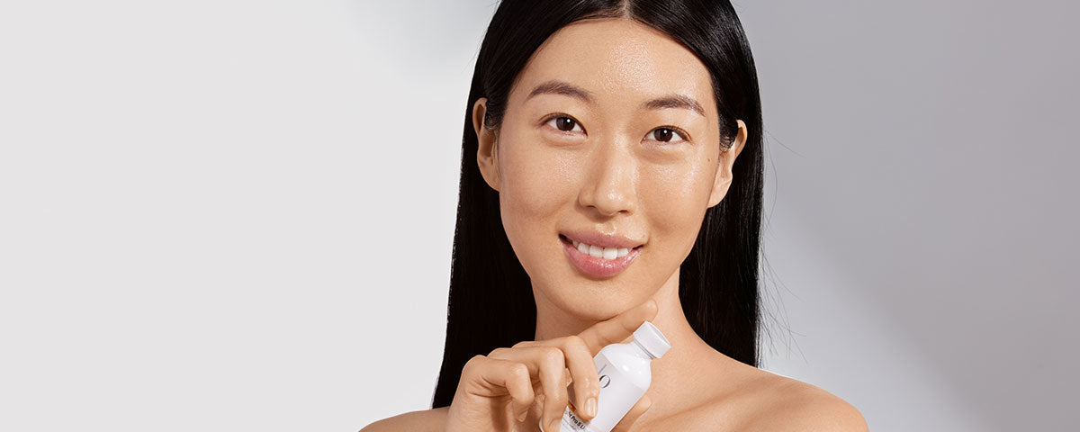 Smiling woman with clear skin holding a white skincare bottle at her shoulder