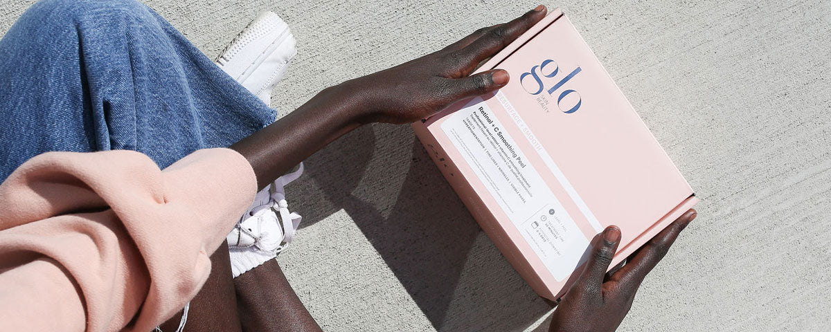 Person holding a pink 'glo' box labeled 'Retinol + Glycolic Peel' while sitting on concrete, wearing jeans and white sneakers.