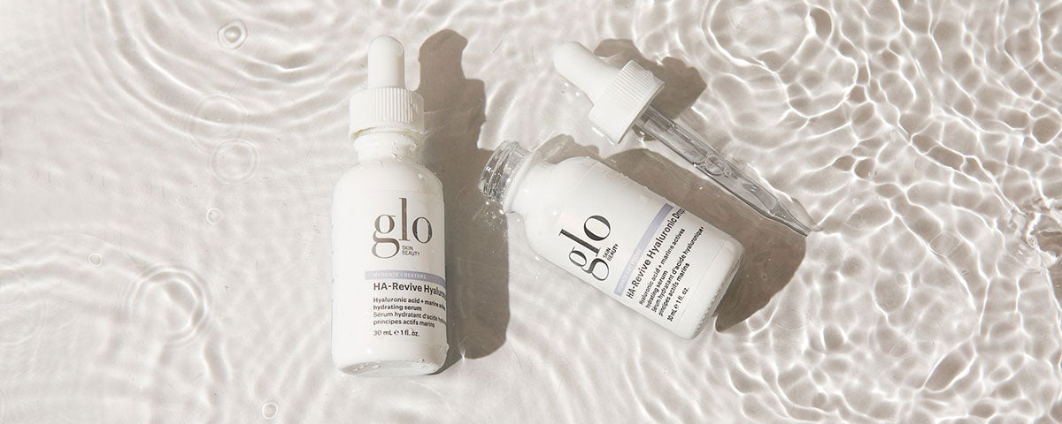 Two glo HA-Revive Hyaluronic Hydrating serum bottles and dropper on rippled water background — 30 mL e 1 fl. oz.