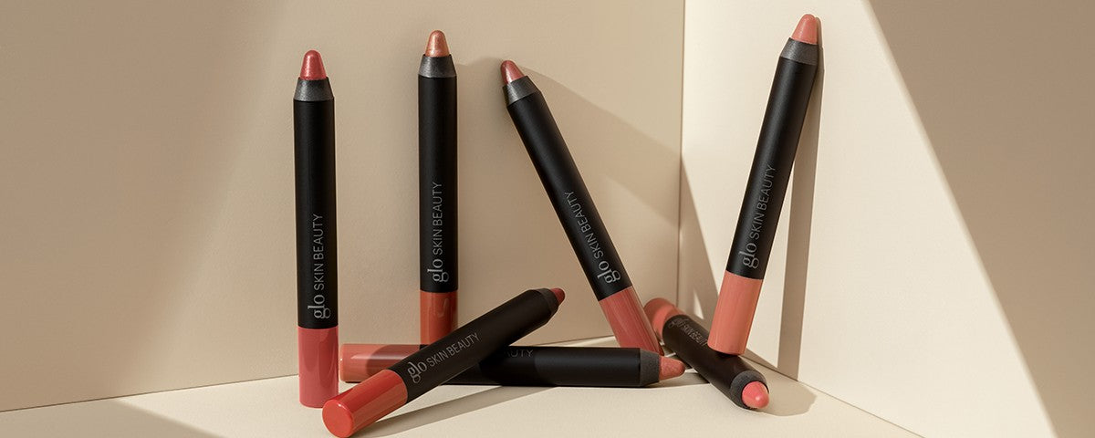 Assorted nude lip crayons on beige surfaces, black tubes labeled 'glo SKIN BEAUTY'.