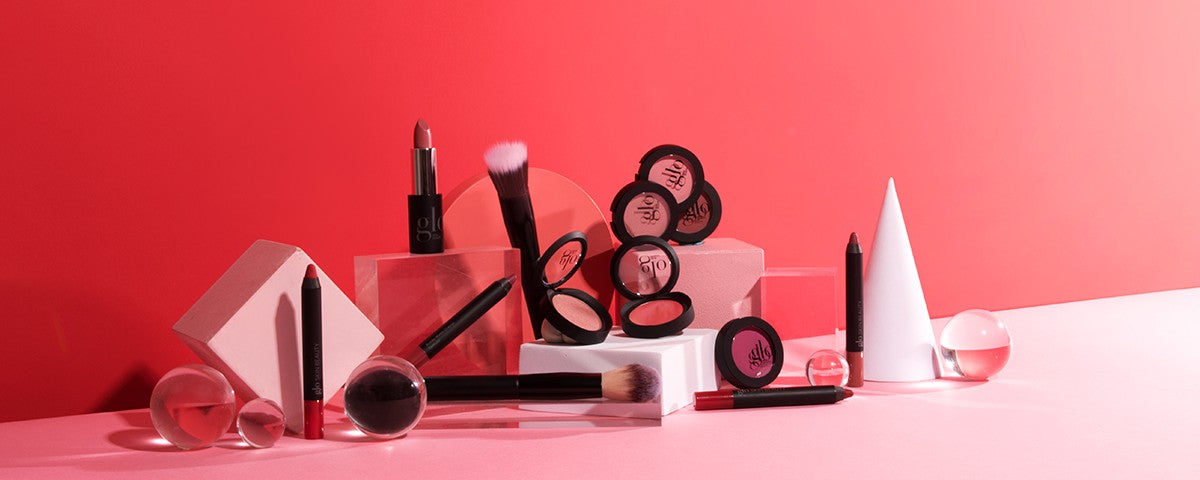 Assorted glo blush compacts, makeup brushes, lipsticks and pencils arranged on pink-red display blocks