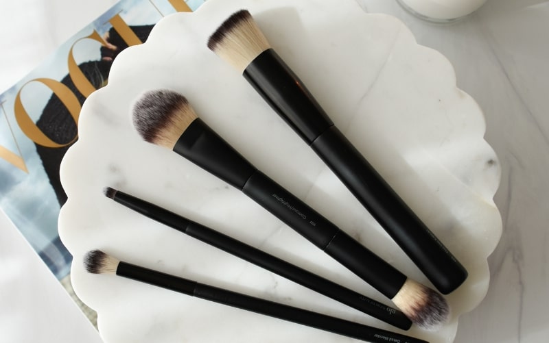 Five black-handled makeup brushes on a scalloped white marble tray over a VOGUE magazine.