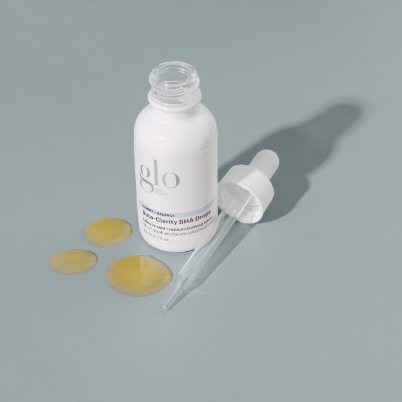 glo skin beauty beta-Clarity BHA Drops bottle with glass dropper and three yellow drops on gray background.