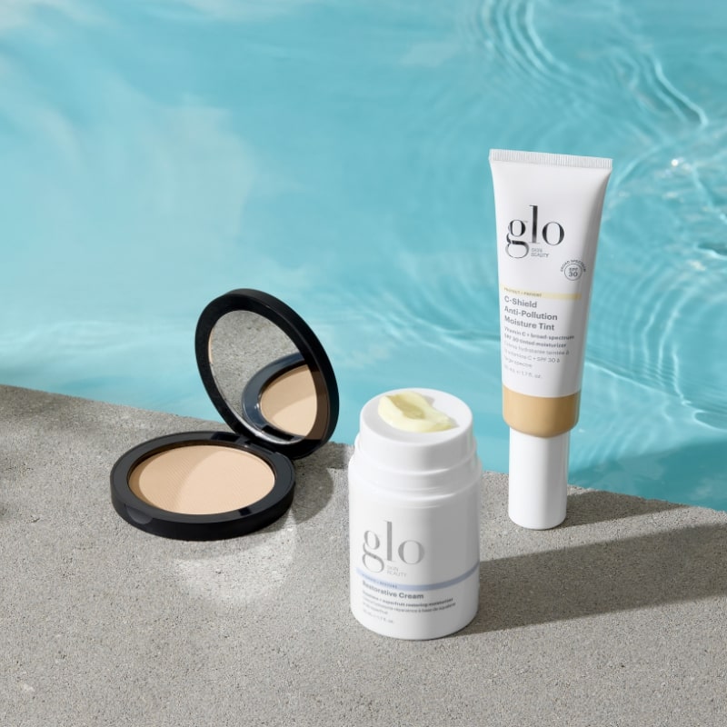 Three glo products by a pool: compact powder, Restorative Cream stick, and C-Shield Anti-Pollution Moisture Tint tube