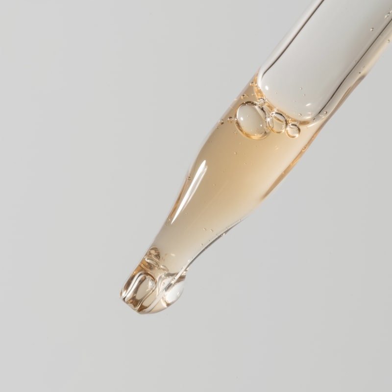 Amber serum droplet at the tip of a glass dropper with tiny bubbles against a light gray background.