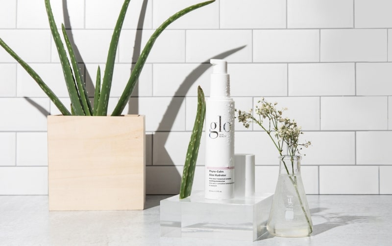 glo Phyto-Calm Aloe Hydrator pump bottle beside an aloe plant and a small beaker with white flowers on tile backdrop