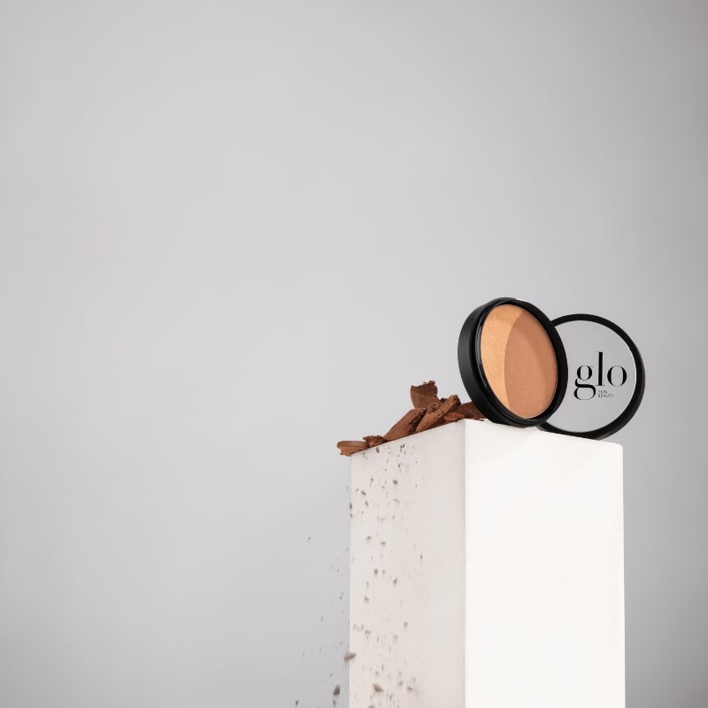 Compact bronzer and highlighter on a white pedestal with brown bark fragments; lid shows text 'glo'.