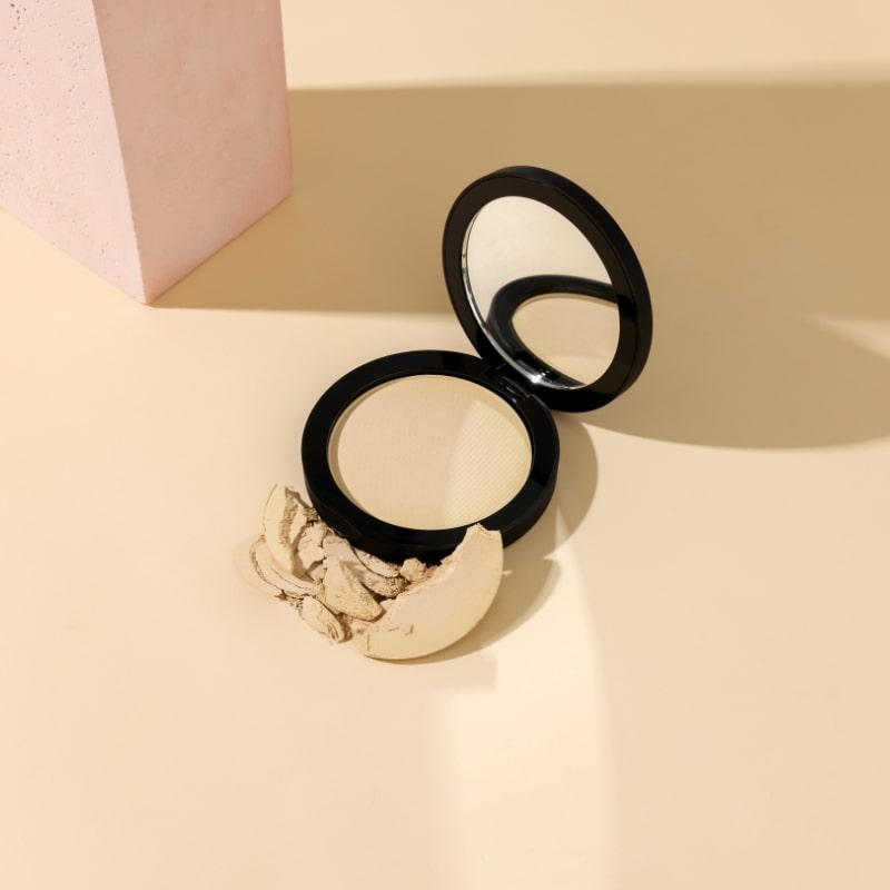 Compact pressed powder in a black compact with mirror and broken powder pieces on a beige surface