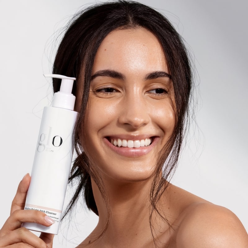 Smiling woman holding a white bottle labeled glo AHA Cleanser next to her face.