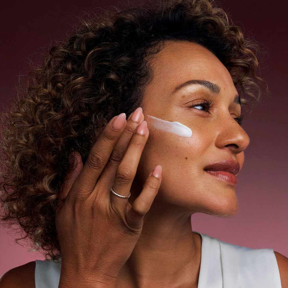 Woman applying Bio-Renew EGF Cream to her cheek against a pink gradient background