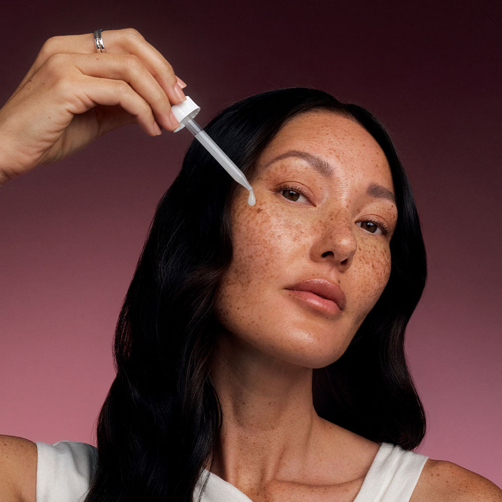 Woman applying Bio-Renew EGF Drops serum with a dropper to her cheek against a pink gradient background