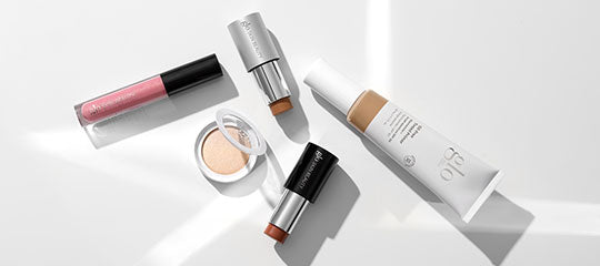Five cosmetics on white surface: lip gloss, pressed powder compact, two stick foundations, and tinted face tube.