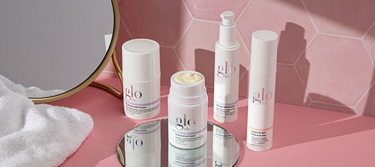 Four white glo skincare bottles and jar arranged on a round mirror with pink tiled backdrop, towel and mirror edge