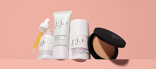 Four glo-branded makeup products on a peach background: dropper bottle, primer tube, pump jar, and open compact powder.