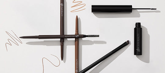 Three brown eyebrow pencils with spoolie, angled brush and brow gel tube arranged on white background.
