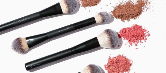Three black-handled makeup brushes with scattered brown and pink powder blush on white background.