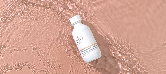 White bottle labeled glo floating in shallow rippled pink-peach water, overhead view