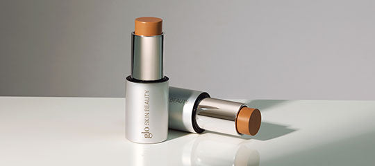 Two glo SKIN BEAUTY cream sticks—one upright, one lying down—showing warm tan shade.