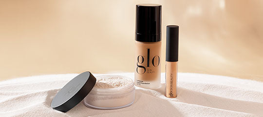 Three glo cosmetic products: open jar of loose powder with black lid, glo foundation bottle, and concealer tube on beige fabric.