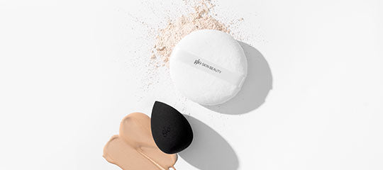 Powder puff labeled 'glo skin beauty', black makeup sponge, beige foundation smear and scattered loose powder.