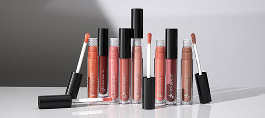 Seven lip gloss tubes in pink and nude shades with open applicators on a reflective surface